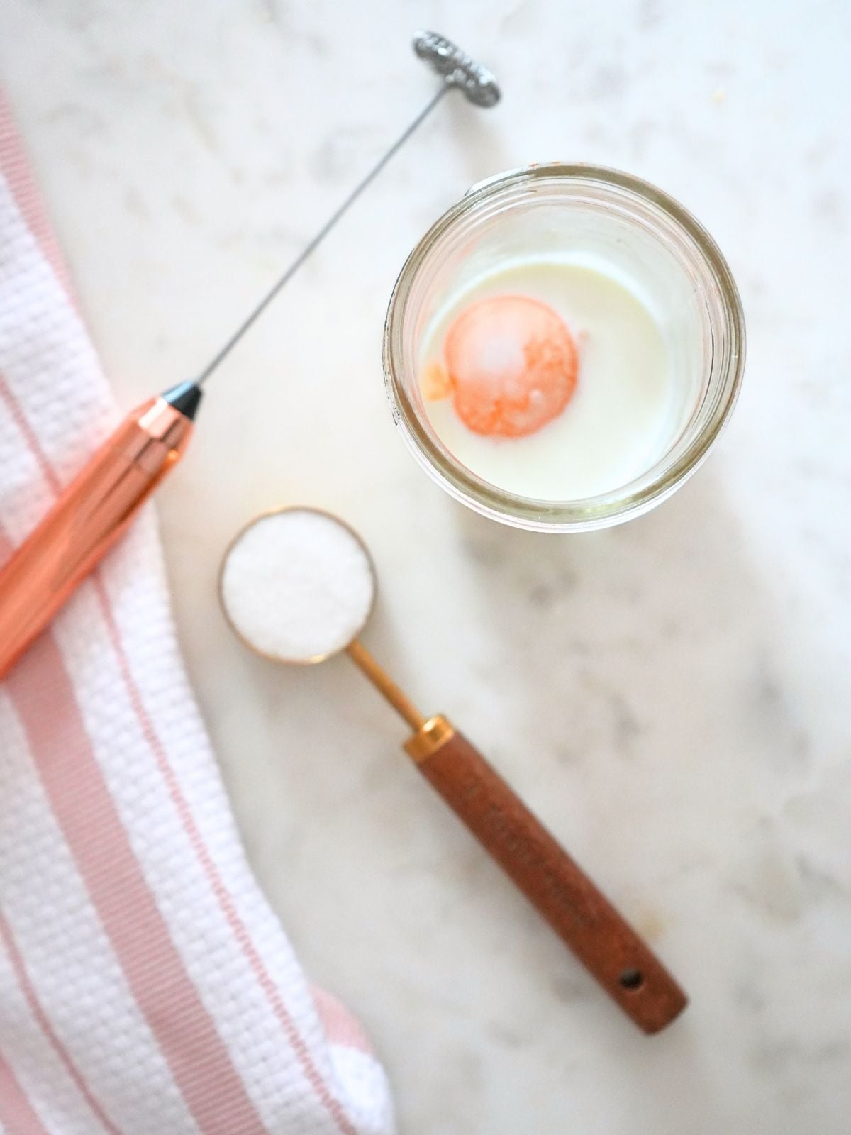 Egg and milk in a jar next to a tablespoon of sugar and a frother.