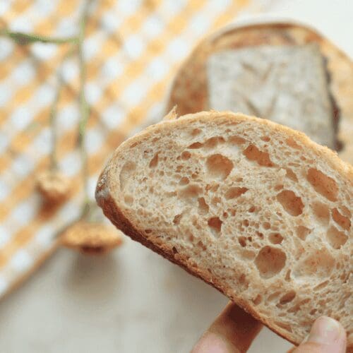 A slice of sourdough bread with a boule and flower in the background