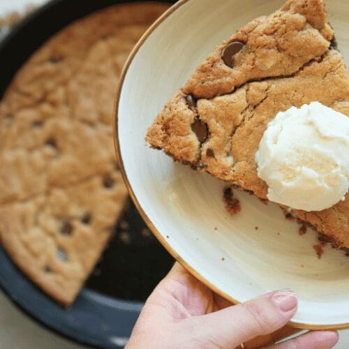 Woman holding slice of sourdough chocolate chip skillet cookie topped with ice cream with the cast iron skillet cookie in the background