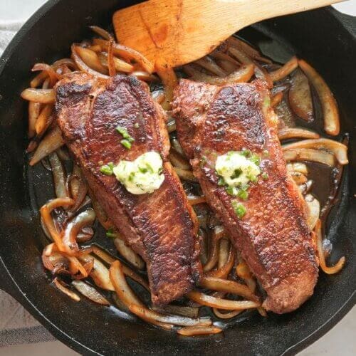 Steaks and onions in cast iron pan with pan sauce and herb butter. Shown with wooden spatula, basil, and decorative pot holder