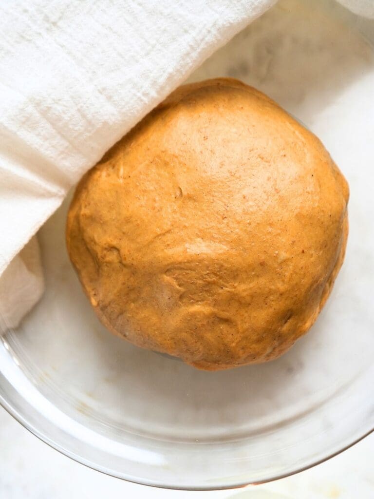 Dough ball in glass bowl partially covered with a linen towel.