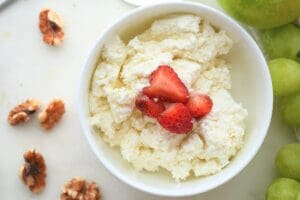 Small bowl of homemade cottage cheese with strawberries on top, shown with walnuts and grapes next to bowl