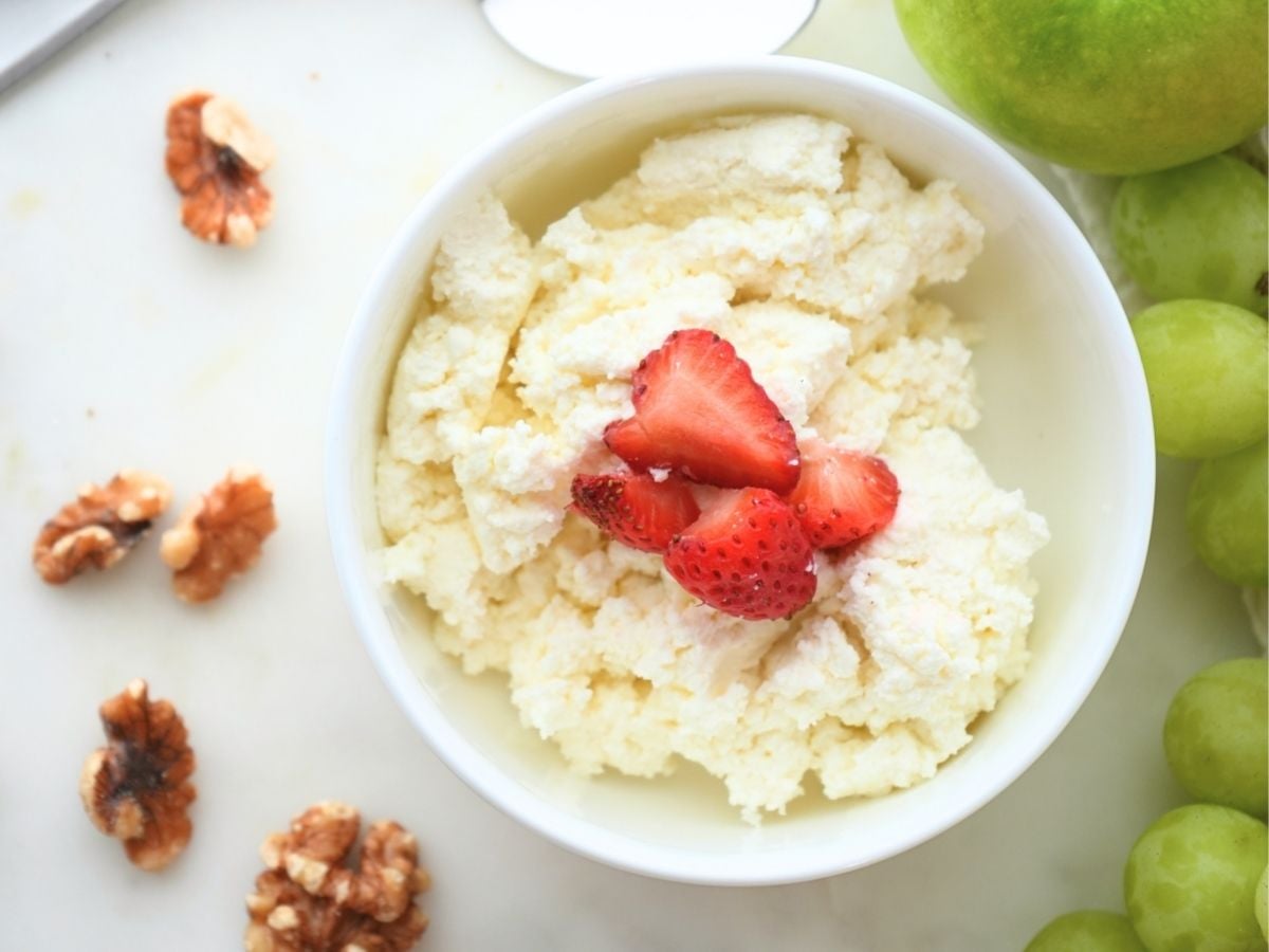 Small bowl of homemade cottage cheese with strawberries on top, shown with walnuts and grapes next to bowl
