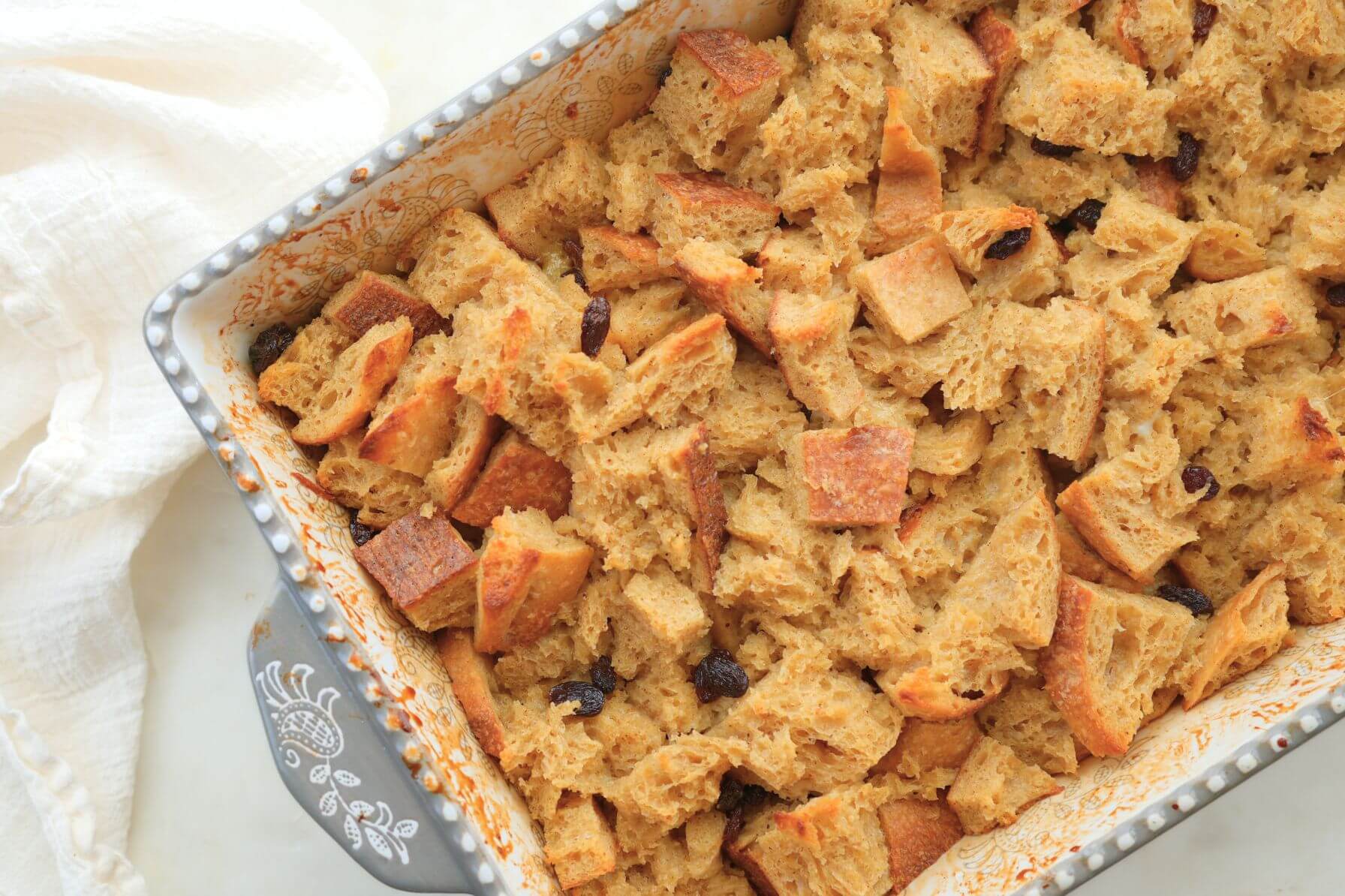 Baked sourdough egg nog bread pudding with golden crust and raisins in a decorative baking dish.