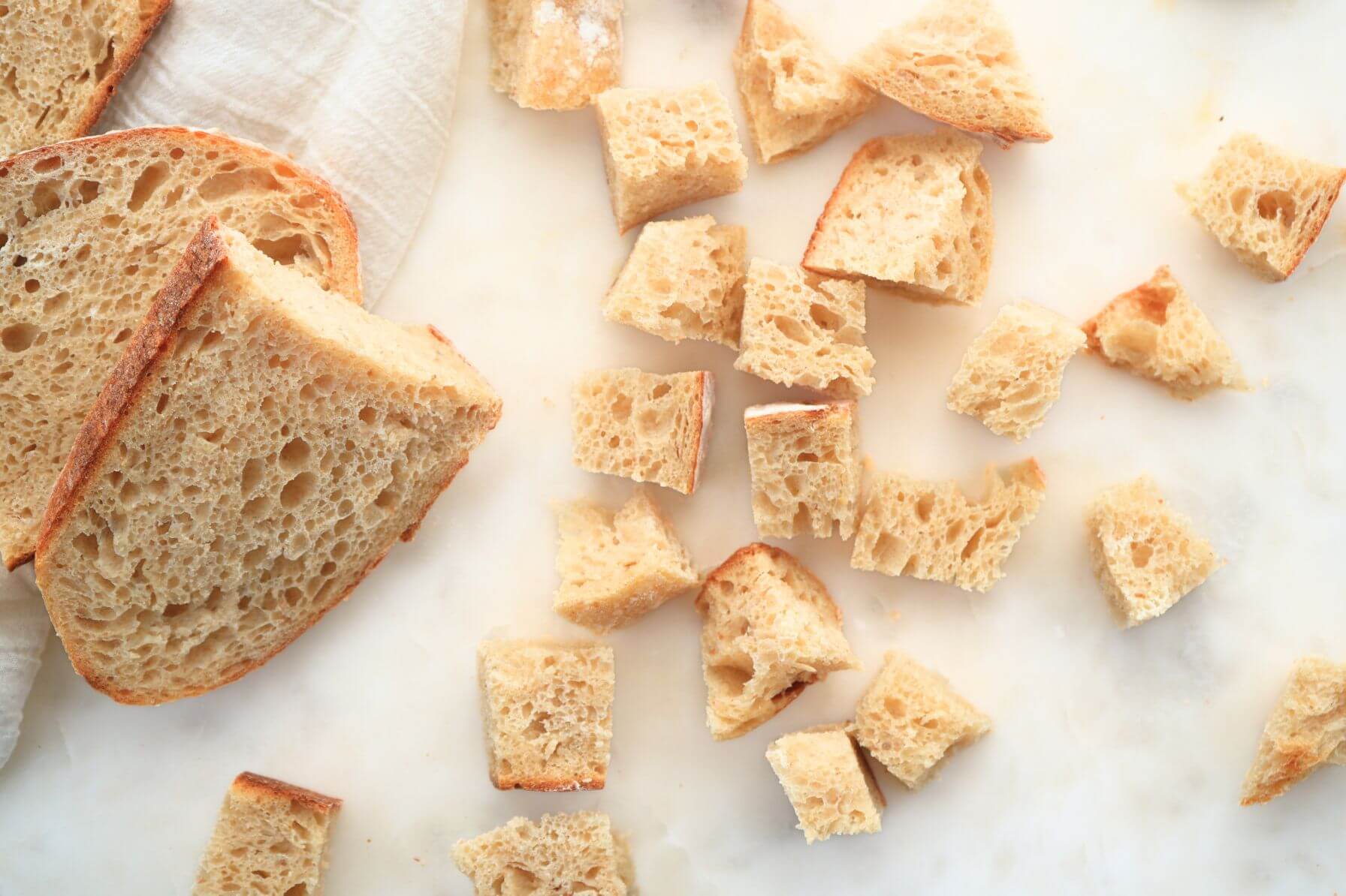 Sliced sourdough bread and cubed pieces arranged on a marble surface with a linen cloth.