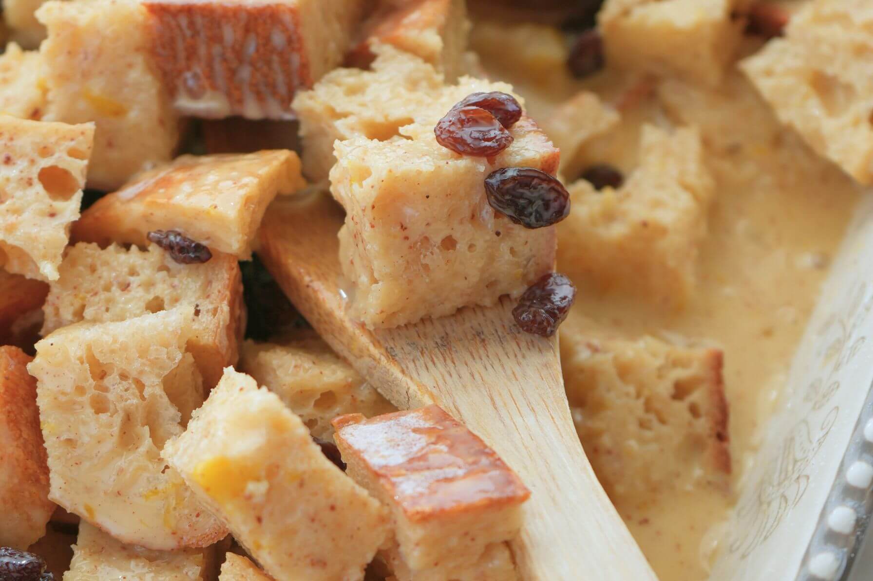 Wooden spoon scooping bread pudding mixture with custard and raisins in a baking dish.