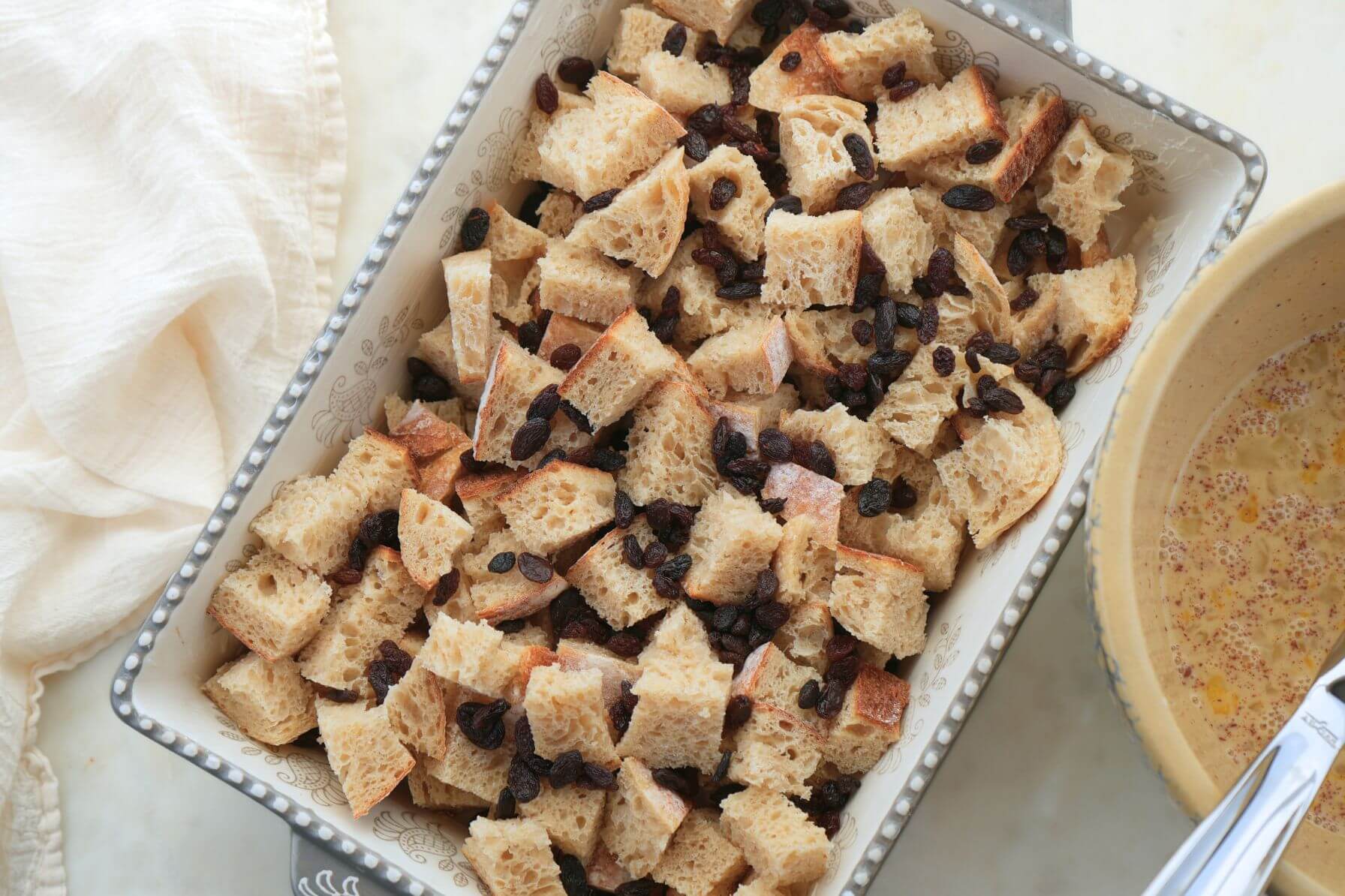 Baking dish filled with sourdough cubes and raisins next to a bowl of egg custard mixture with a whisk.