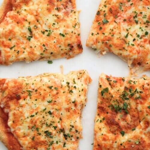 Four slices of cheesy pizza topped with herbs on a white marble surface, surrounded by scattered dried herbs and red pepper flakes.