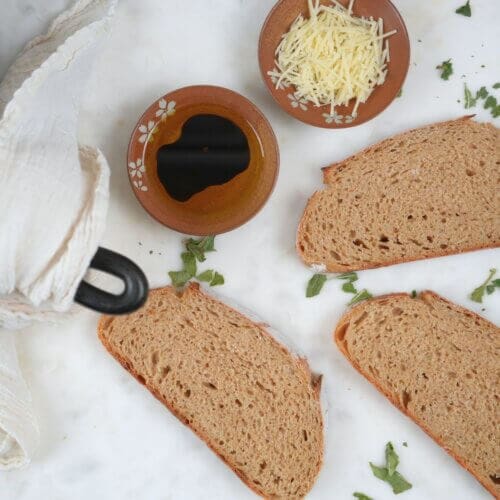 Freshly sliced sourdough bread made with fresh-milled flour, served with balsamic dip, shredded cheese, and garnished with fresh herbs on a marble surface.