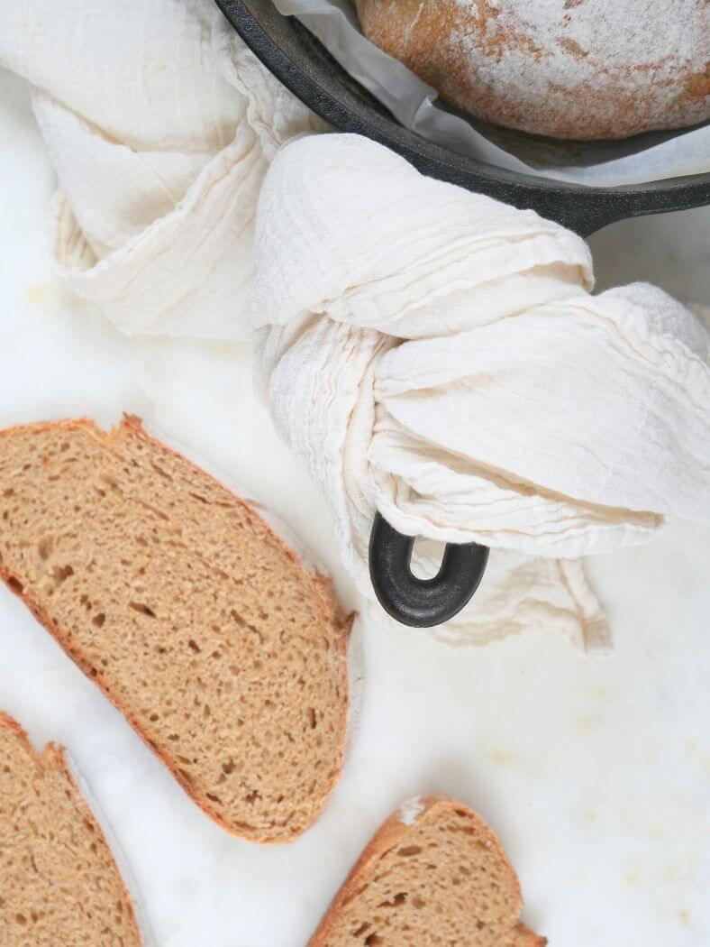 Fresh-milled sourdough bread with slices on a white surface and a cast iron pot in the background