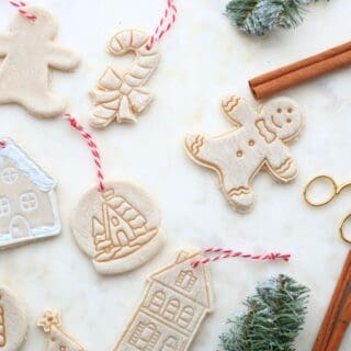Handmade salt dough ornaments shaped like gingerbread men, candy canes, and houses, displayed with cinnamon sticks, twine, gold scissors, and snowy evergreen sprigs on a white marble background.
