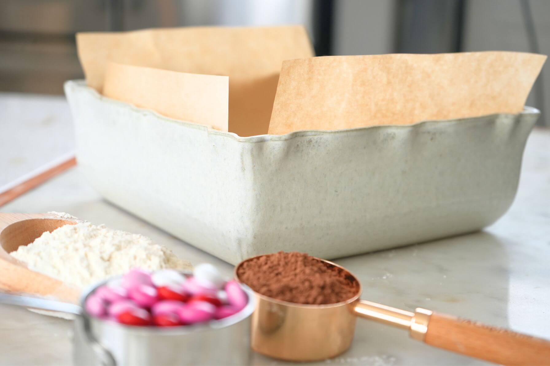 A ceramic baking dish lined with parchment paper with measuring cups of flour, m&m's, and cocoa powder.