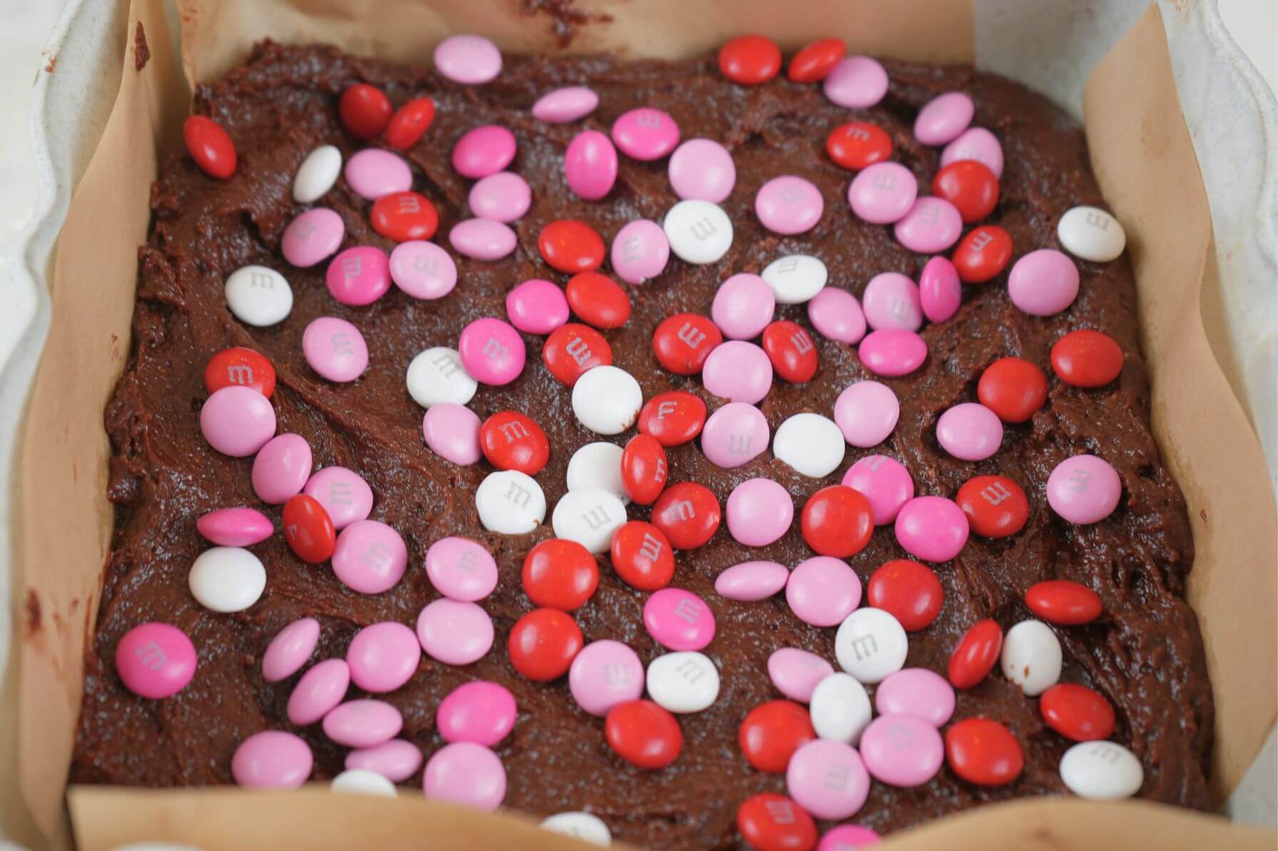 Brownie batter spread in a parchment-lined baking dish, topped with pink, red, and white M&M's.