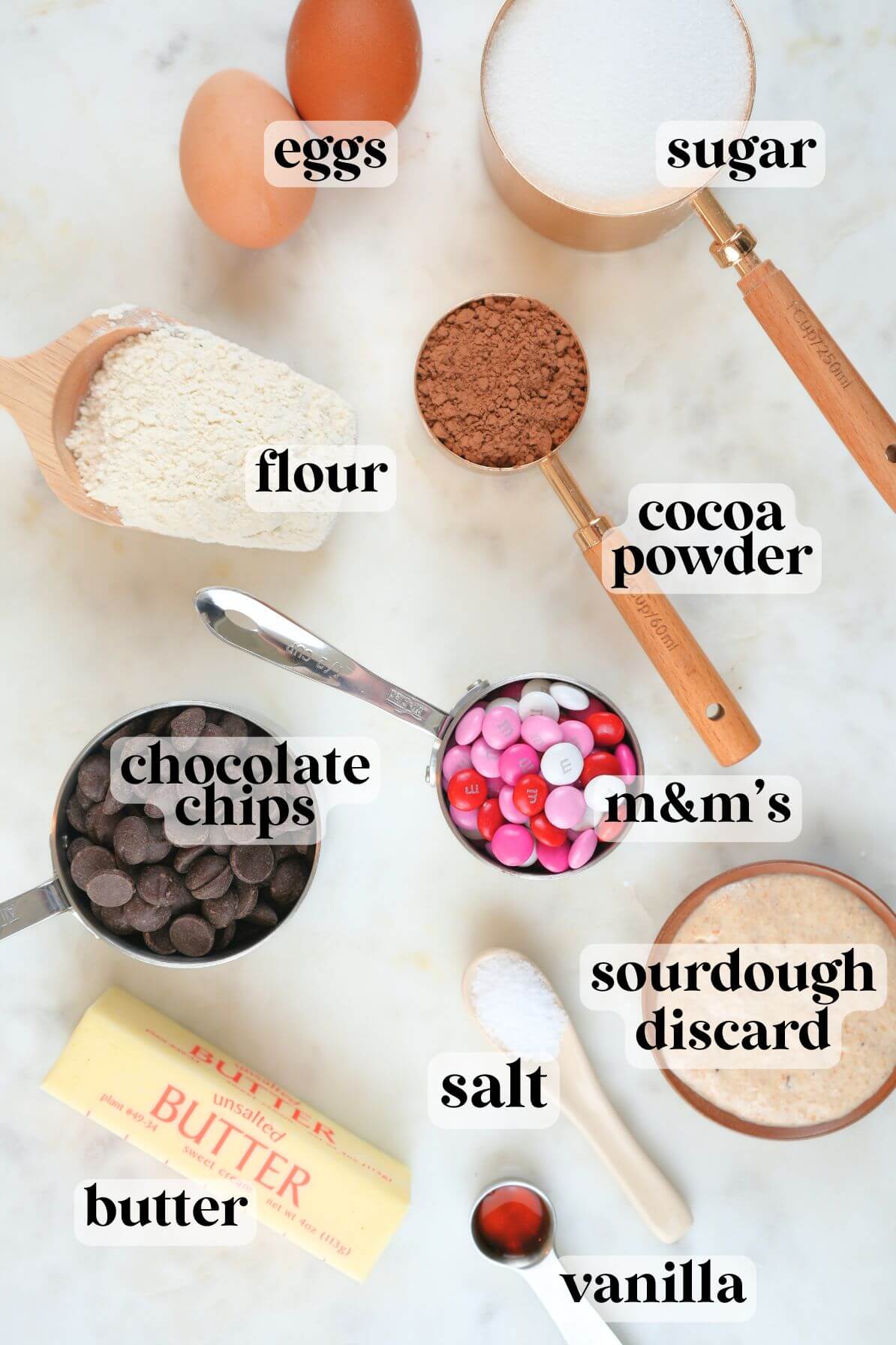 Overhead view of labeled ingredients for sourdough Valentine's brownies, including eggs, flour, sugar, cocoa powder, chocolate chips, M&Ms, sourdough discard, butter, salt, and vanilla