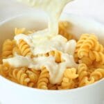 Creamy béchamel sauce being poured over a bowl of rotini pasta.