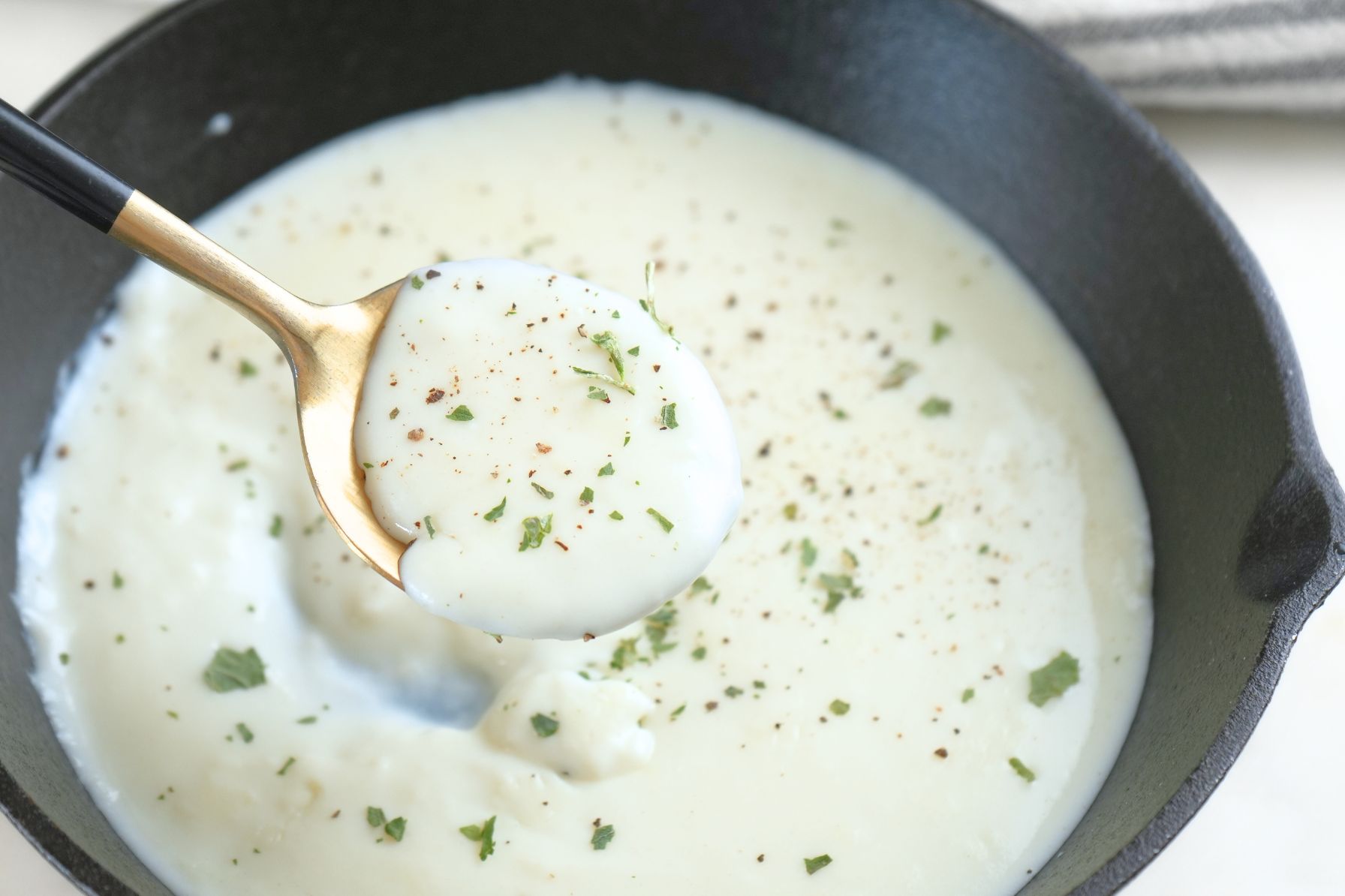 A spoonful of creamy béchamel sauce garnished with black pepper and fresh herbs, lifted from a cast iron skillet.