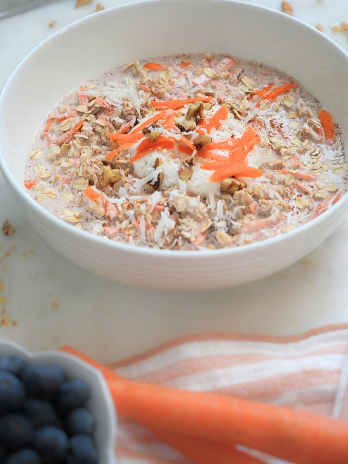 Carrot cake overnight oats in a white bowl with blueberries and whole carrots in the foreground.