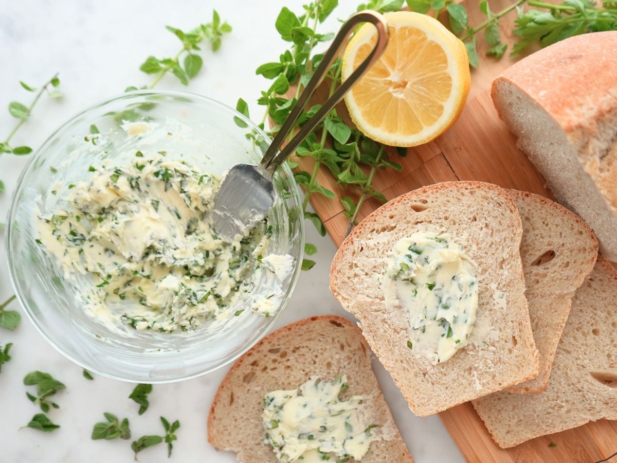 Herb butter in a bowl and on bread, next to a fresh lemon and herbs.