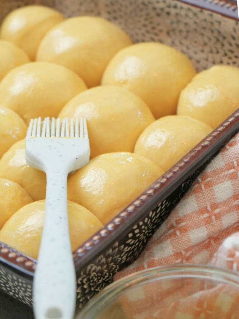 A silicone pastry brush resting on shaped rolls covered in a light egg wash.