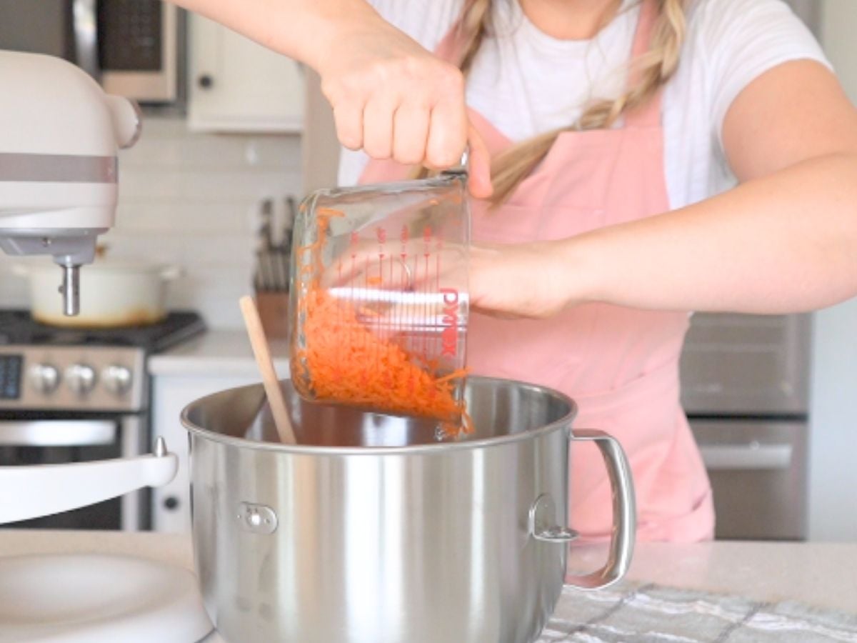 Woman adding shredded carrots to a stand mixer bowl.