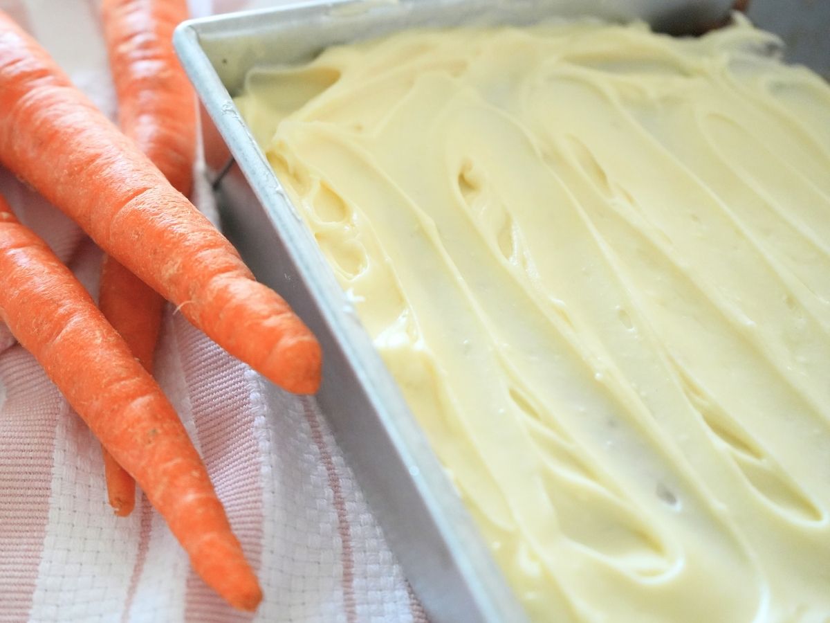 Frosted sourdough carrot cake with fresh carrots on counter.