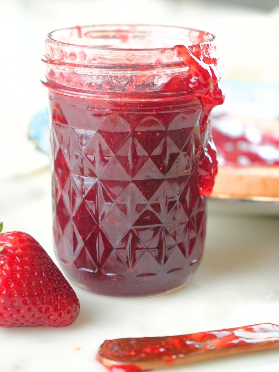 A mason jar filled with strawberry jam and some spilling over the edge, shown with fresh berrie and a gold spreading knife