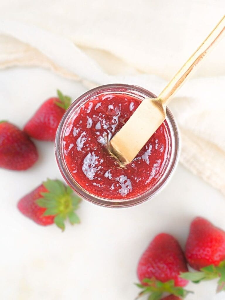 An overhead view of a jar filled with canned strawberry jam with fresh berries scattered along the side