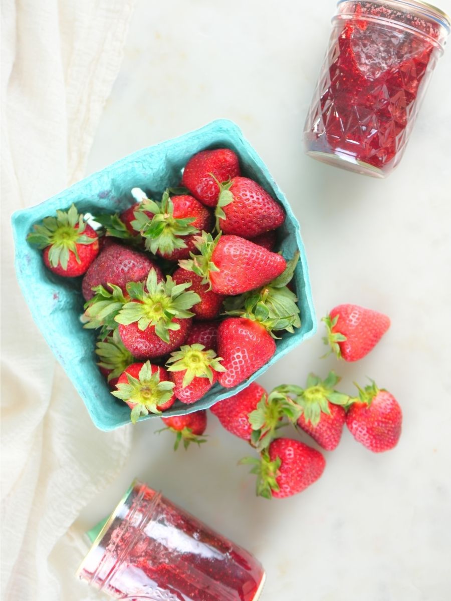 An overhead view of fresh berries and jars of strawberry jam scattered on a white counter