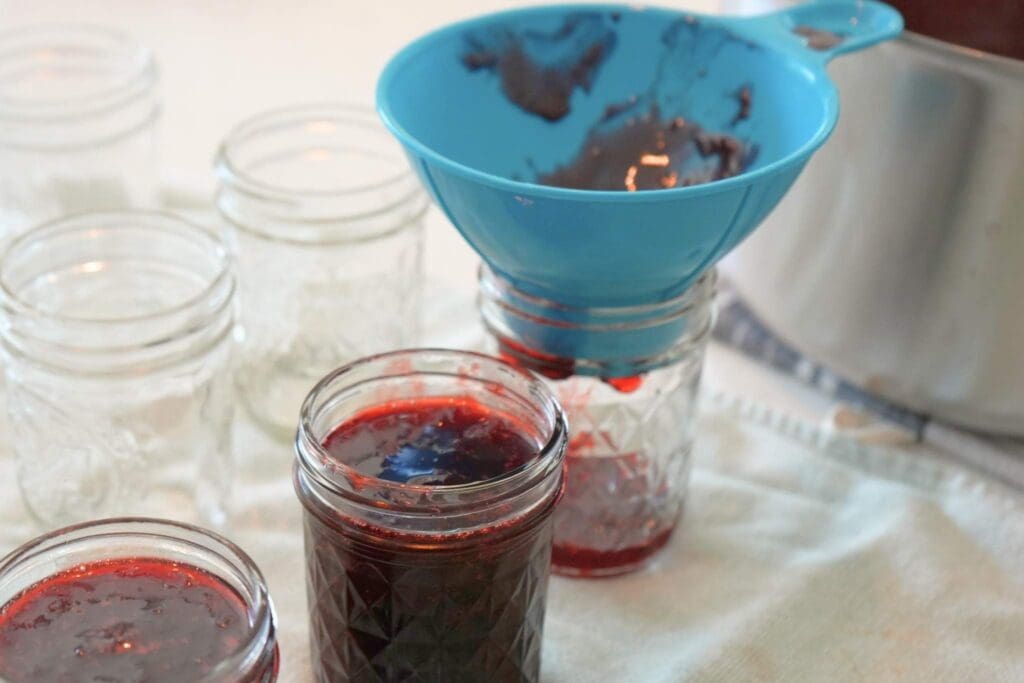 Half pint jars being filled with strawberry jam, one with a blue funnel in it