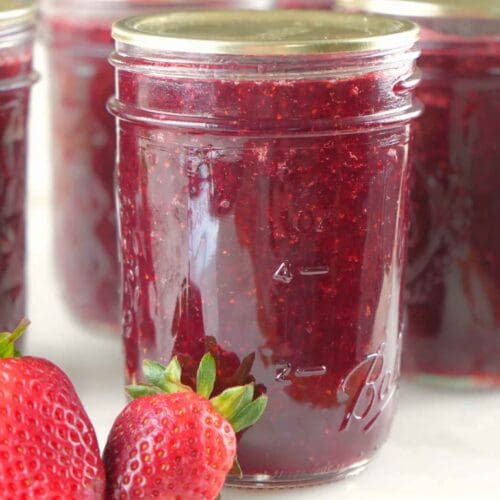 Jars of canned strawberry jam on a counter with fresh strawberries in the foreground