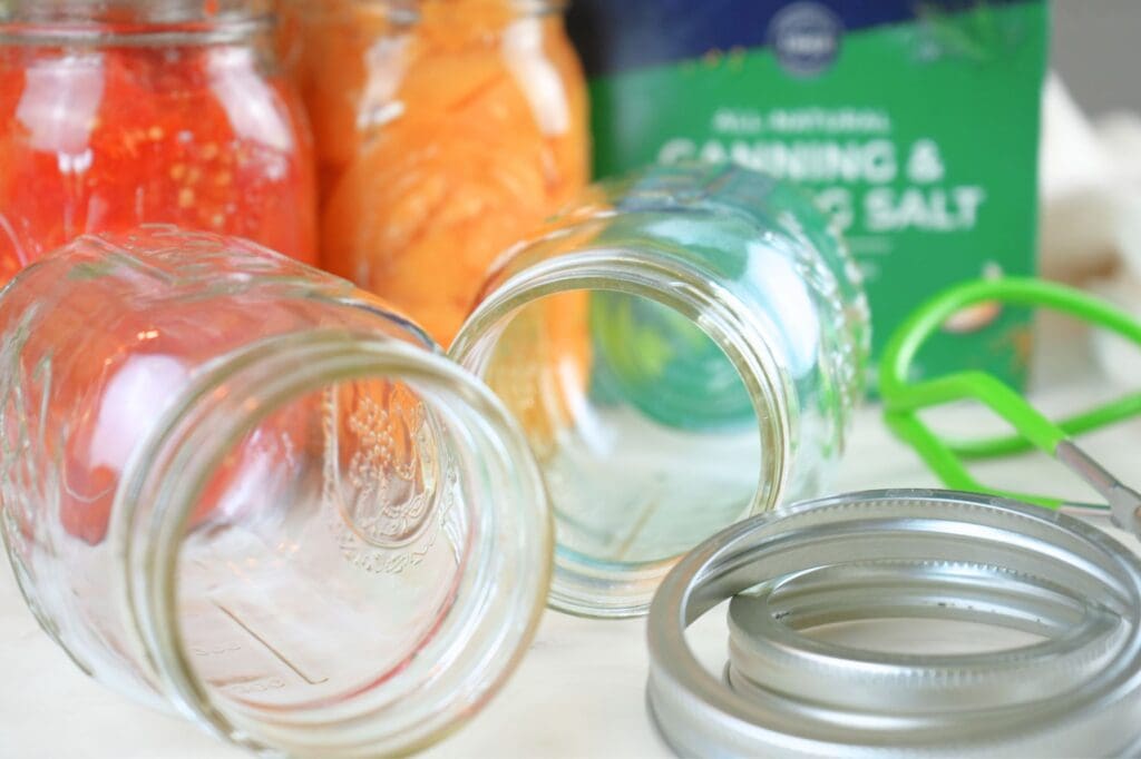 Canning equipment displayed (jars and lids) with home-canned tomatoes, peaches, and a box of canning salt in the background