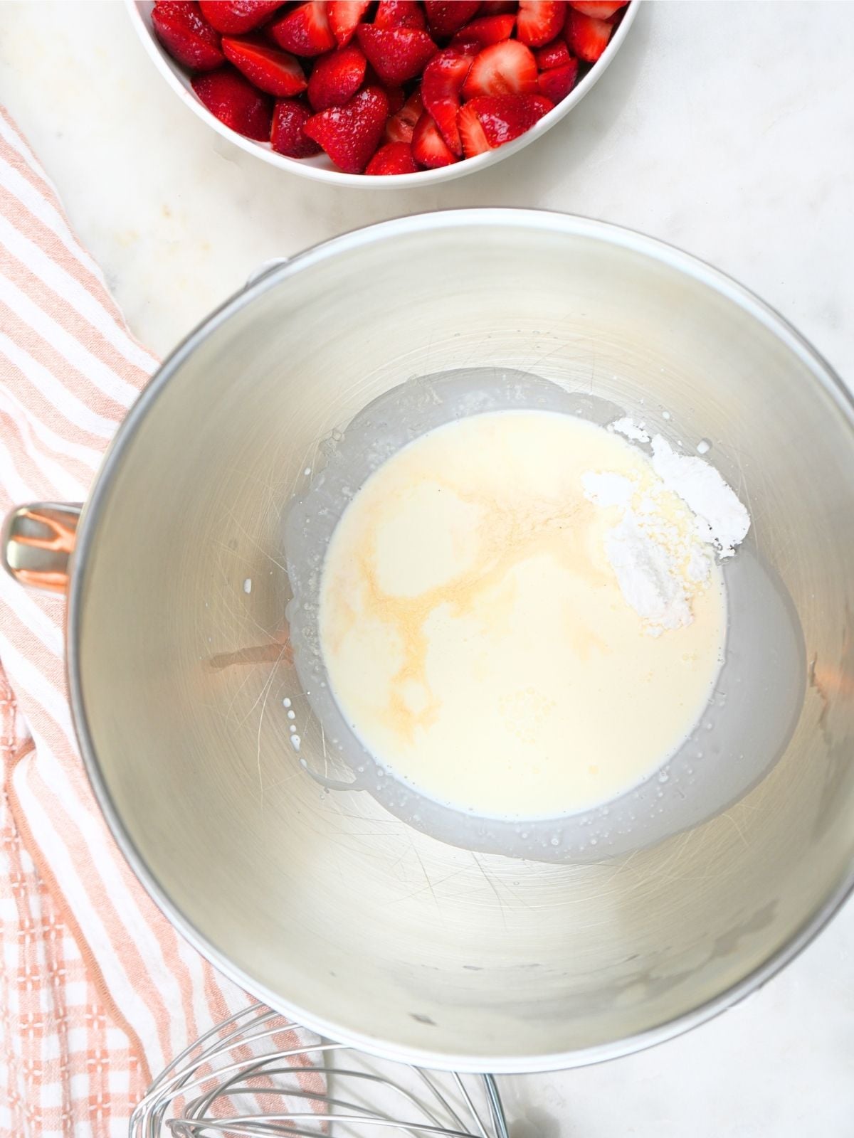 Cream, vanilla, and powdered sugar in a stand mixer bowl, shown with a wire whisk attachment.