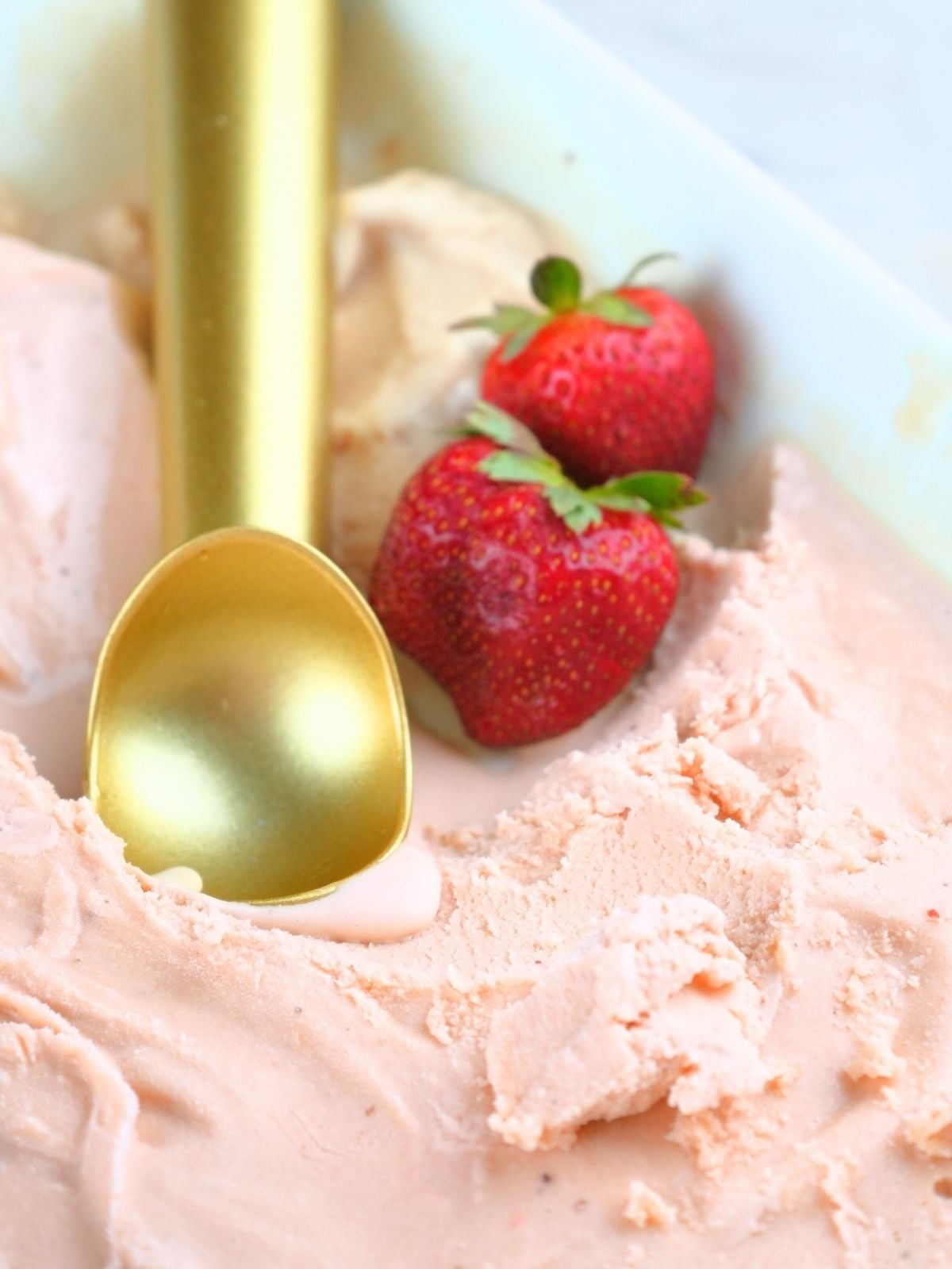A dish of strawberry ice cream with a gold scoop and two fresh berries resting on top