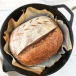 A half fresh-milled sourdough boule on parchment paper in a cast iron Dutch oven, next to a sprig of herbs