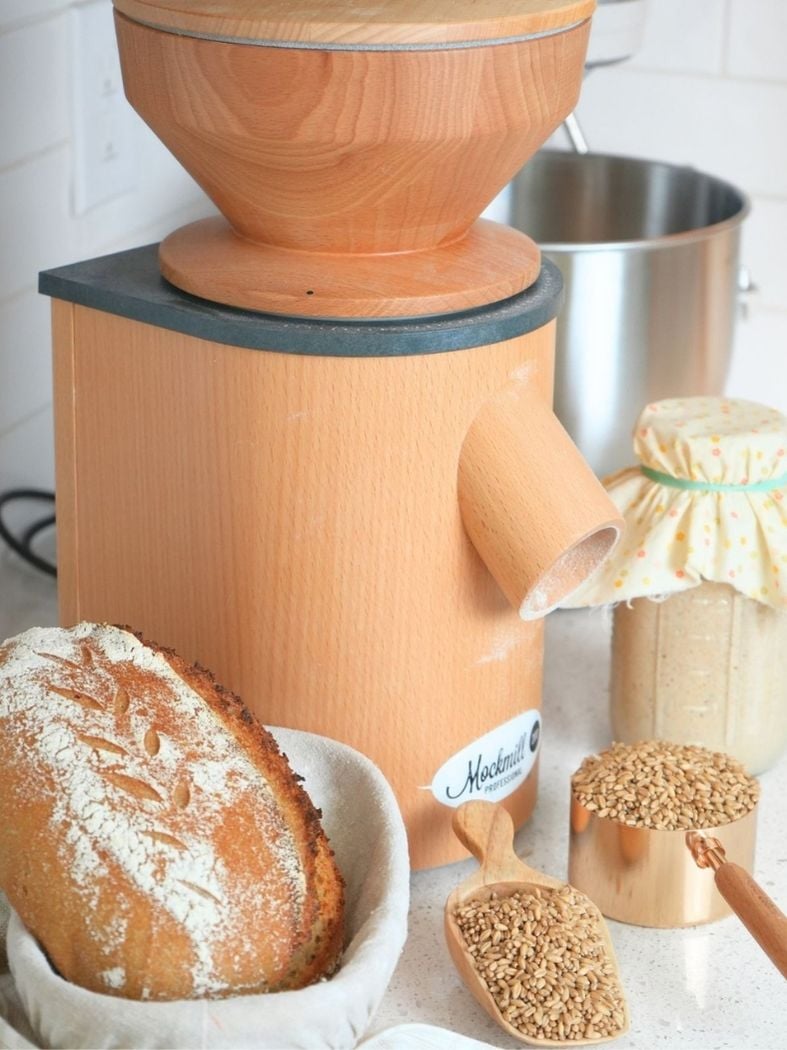 A grain mill with a loaf of sourdough bread, wheat berries, and sourdough starter in the foreground