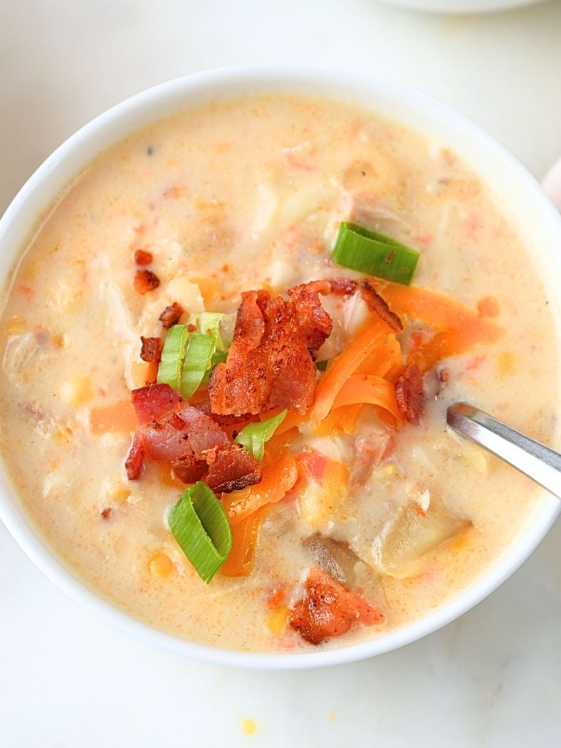 A small white bowl of chicken corn chowder garnished with bacon, green onions, and shredded cheese