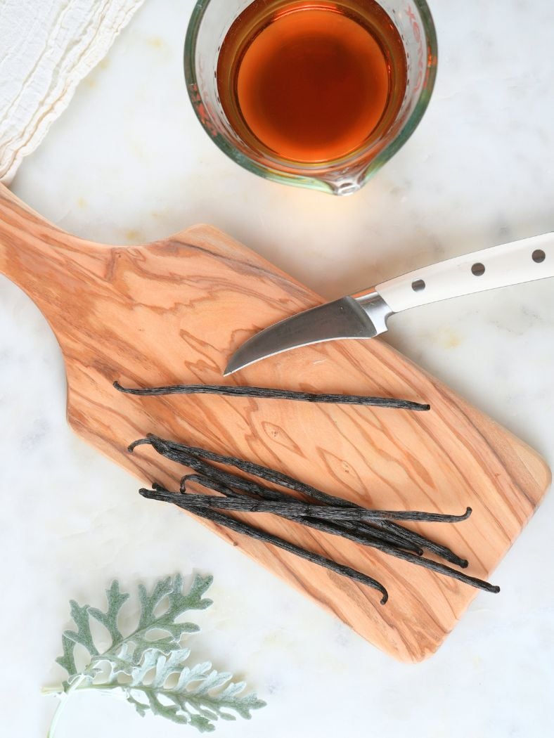 Vanilla beans on a cutting board with a paring knife, shown with bourbon and decorative foliage