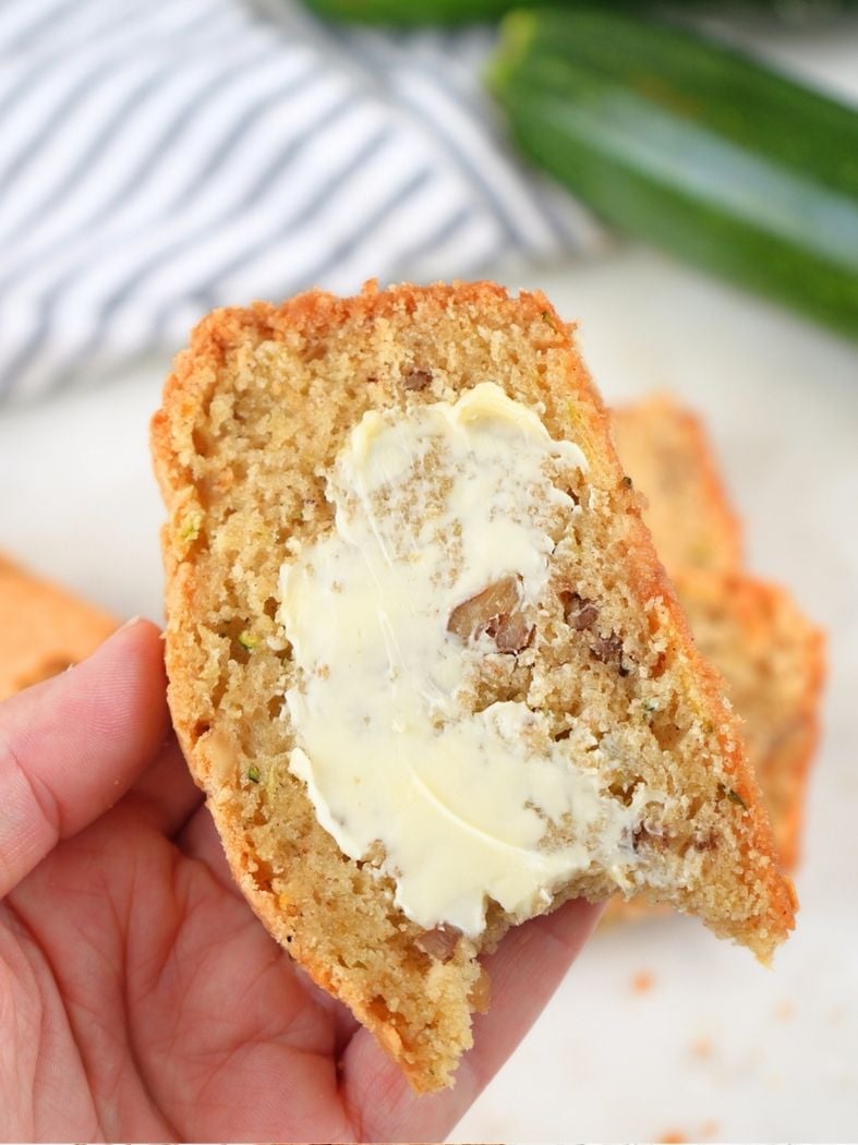 A hand holding a slice of sourdough zucchini bread with a smear of butter and a bite taken out