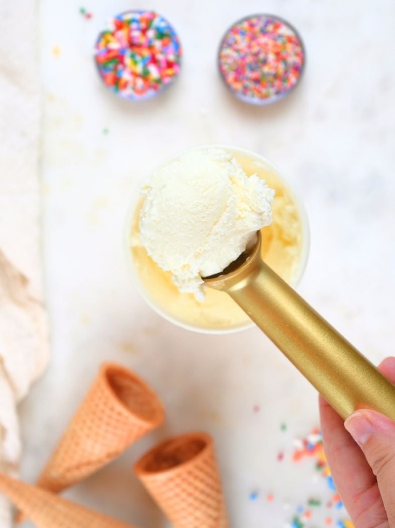 Overhead view of a ice cream scoop with vanilla ice cream with cones and sprinkles in the background