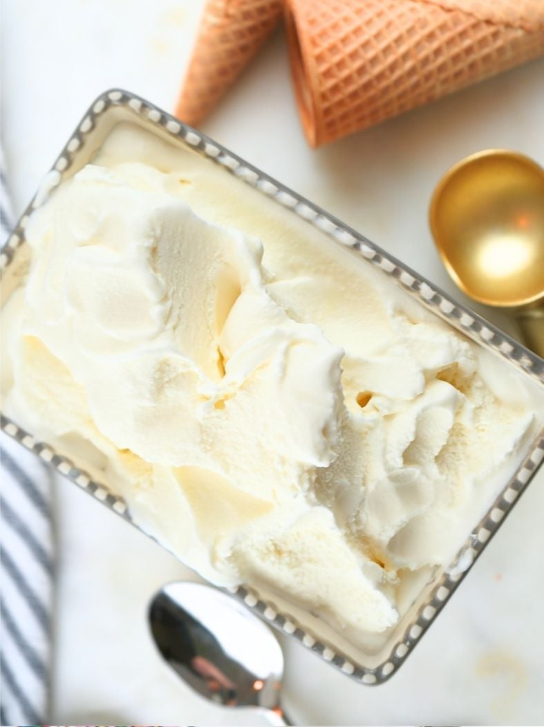 A dish filled with homemade vanilla ice cream (no egg), next to a spoon, scoop, and cones