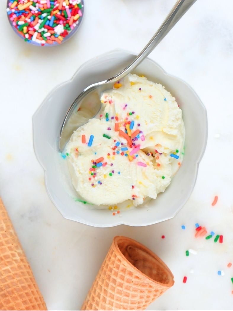 A scoop of eggless vanilla ice cream with sprinkles on top and cones in the background