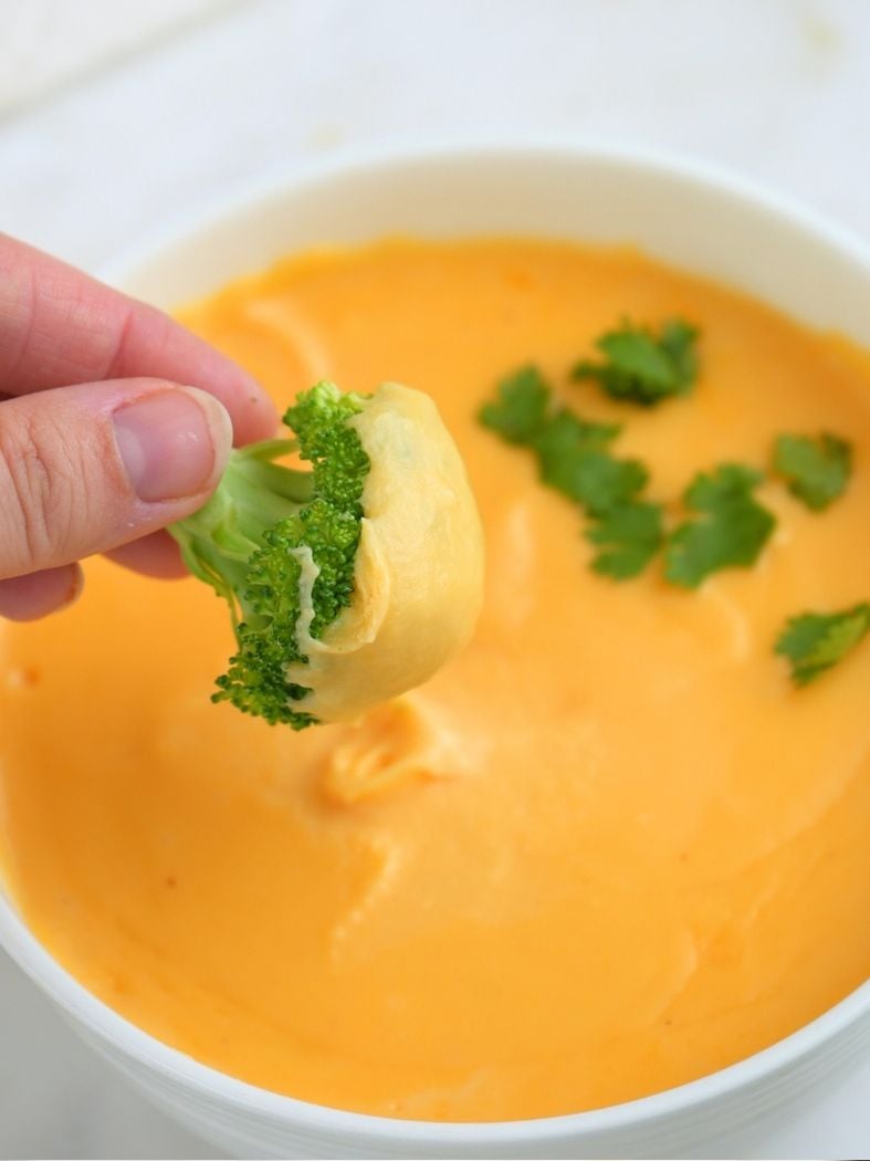 A hand dipping broccoli in a bowl of sourdough discard cheese sauce