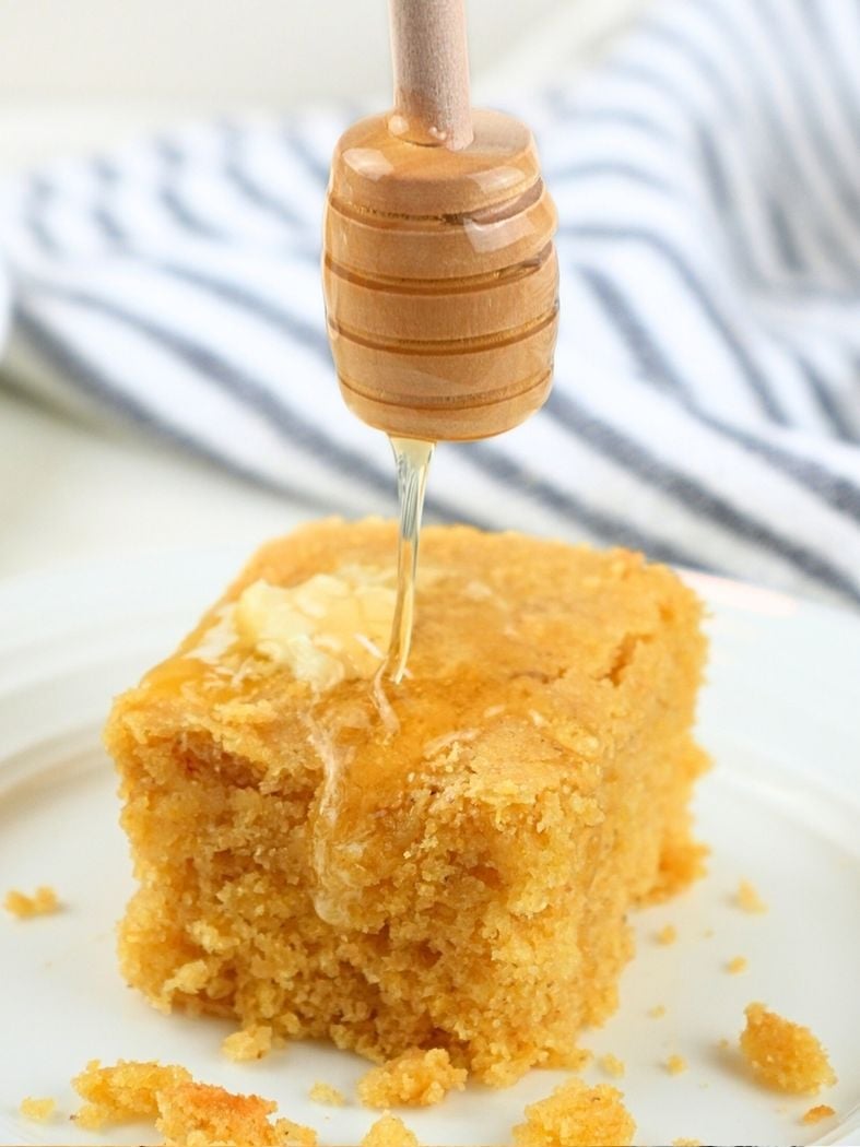 A slice of fresh-milled cornbread with butter on top and honey dipper drizzling honey on top