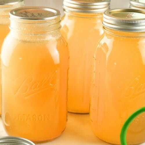 Jars of home-canned chicken broth on a counter with lids and canning tongs in the foreground