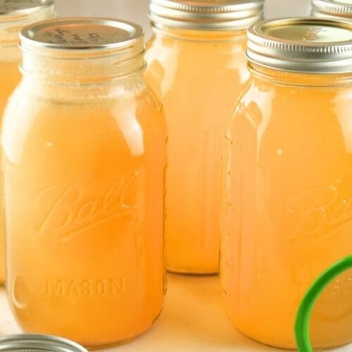 Jars of home-canned chicken broth on a counter with lids and canning tongs in the foreground