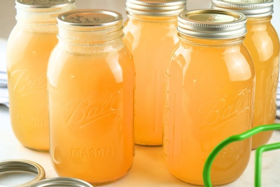 Jars of home-canned chicken broth on a counter with lids and canning tongs in the foreground