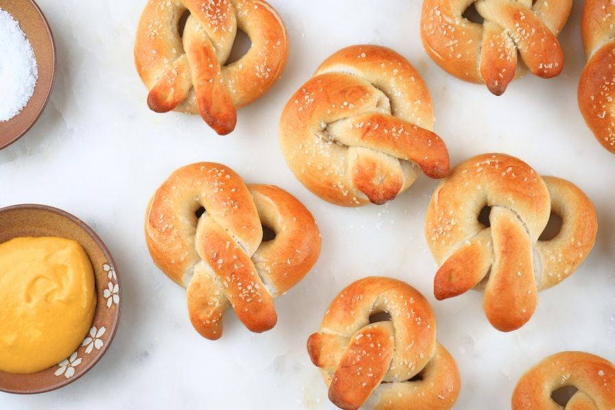 Sourdough soft pretzels scattered on a counter next to a bowls of salt and mustard