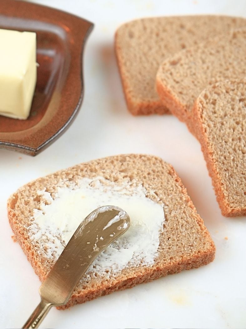 A slice of bread with butter and a gold butter knife, more slices and a dish of butter in the background