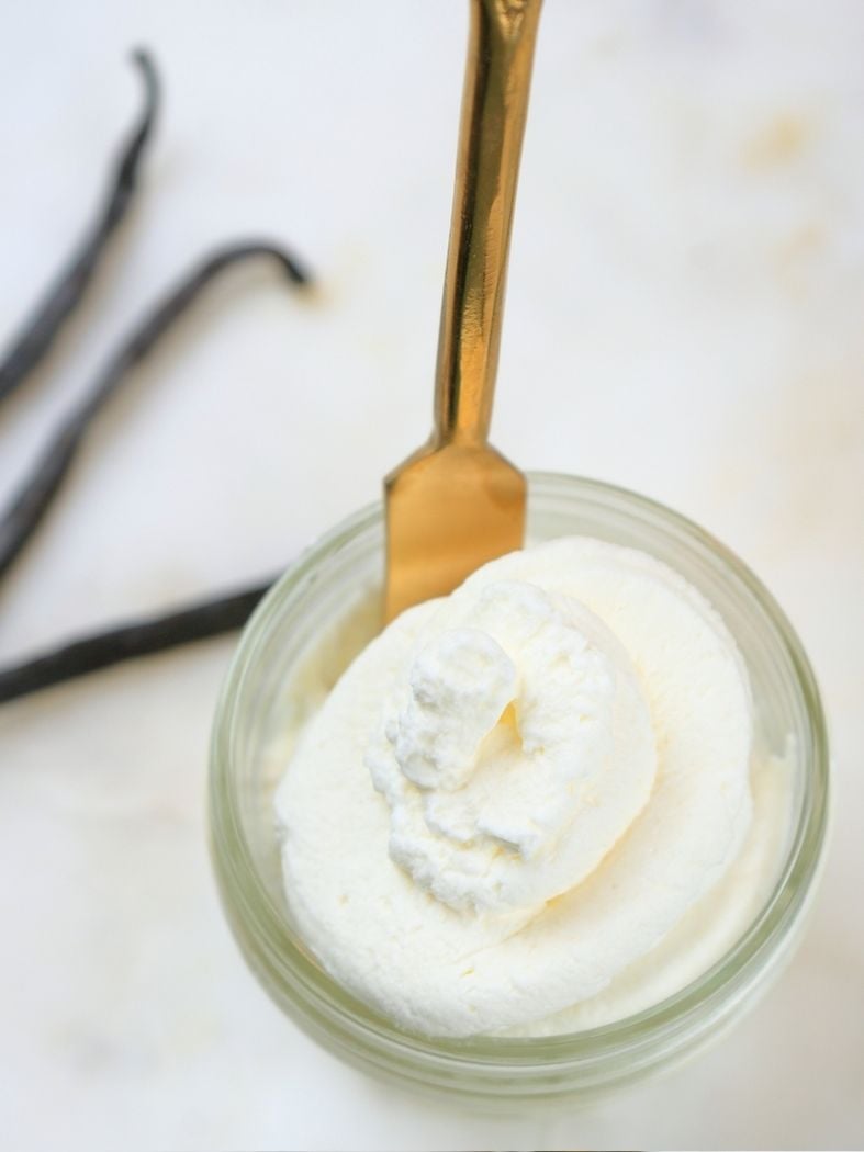 A small bowl of vanilla whipped cream with a gold spoon in it and vanilla beans in the background