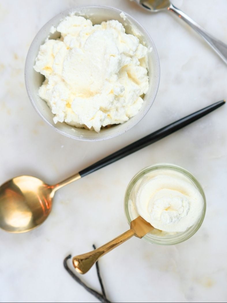 An overhead view of two bowls of homemade vanilla whipped cream, a gold spoon, and vanilla beans